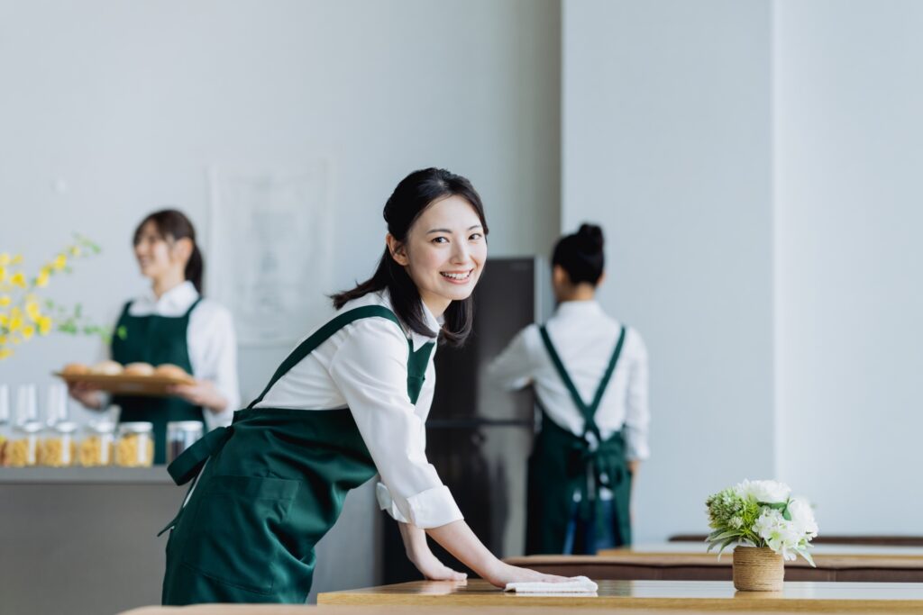 初バイトにおすすめのバイト先記事のアイキャッチ。初めてでも安心感のあるカフェで、笑顔でテーブルを拭く女性スタッフ。